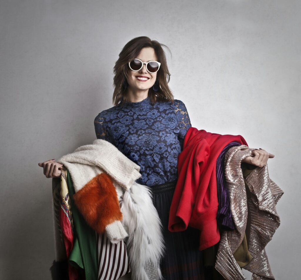Donna sorridente con occhiali da sole che tiene in mano diversi capi di abbigliamento colorati, simbolo dello shopping sfrenato e dell’acquisto compulsivo.