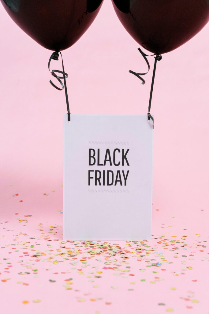 Cartello con la scritta “Black Friday” appeso a due palloncini neri su sfondo rosa con coriandoli colorati.
